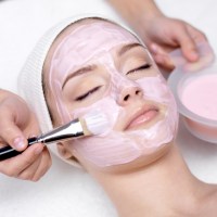 A woman receiving a professional facial with a pink mask being applied with a mask brush